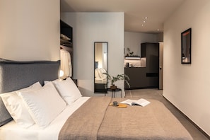 Ein modernes Hotelzimmer mit einem ordentlich bezogenen Bett, einem Nachttisch mit einem Buch und einer Tasse, einem Spiegel sowie Blick ins Bad und die Küchenzeile.
