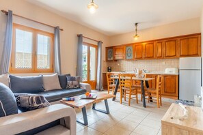 Ein modernes Wohnzimmer mit Sofa und Couchtisch, eine Küche mit Holzschränken und einem Kühlschrank, sowie große Fenster mit Vorhängen.