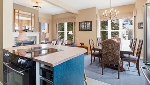 A modern kitchen with a central island, a dining area with a large table and chairs, and a view of the outdoors through large windows. - Fabulous 5-bedroom house at Silver Star Mountain perfect for family getaways! (Vernon)