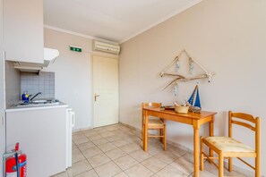 Eine kleine Küche mit einem weißen Kühlschrank, einem Herd und einem Spülbecken. Auf dem Boden steht ein Feuerlöscher. Ein Holztisch mit Stühlen lehnt an der Wand, darüber hängt ein kleines dekoratives Segelboot.