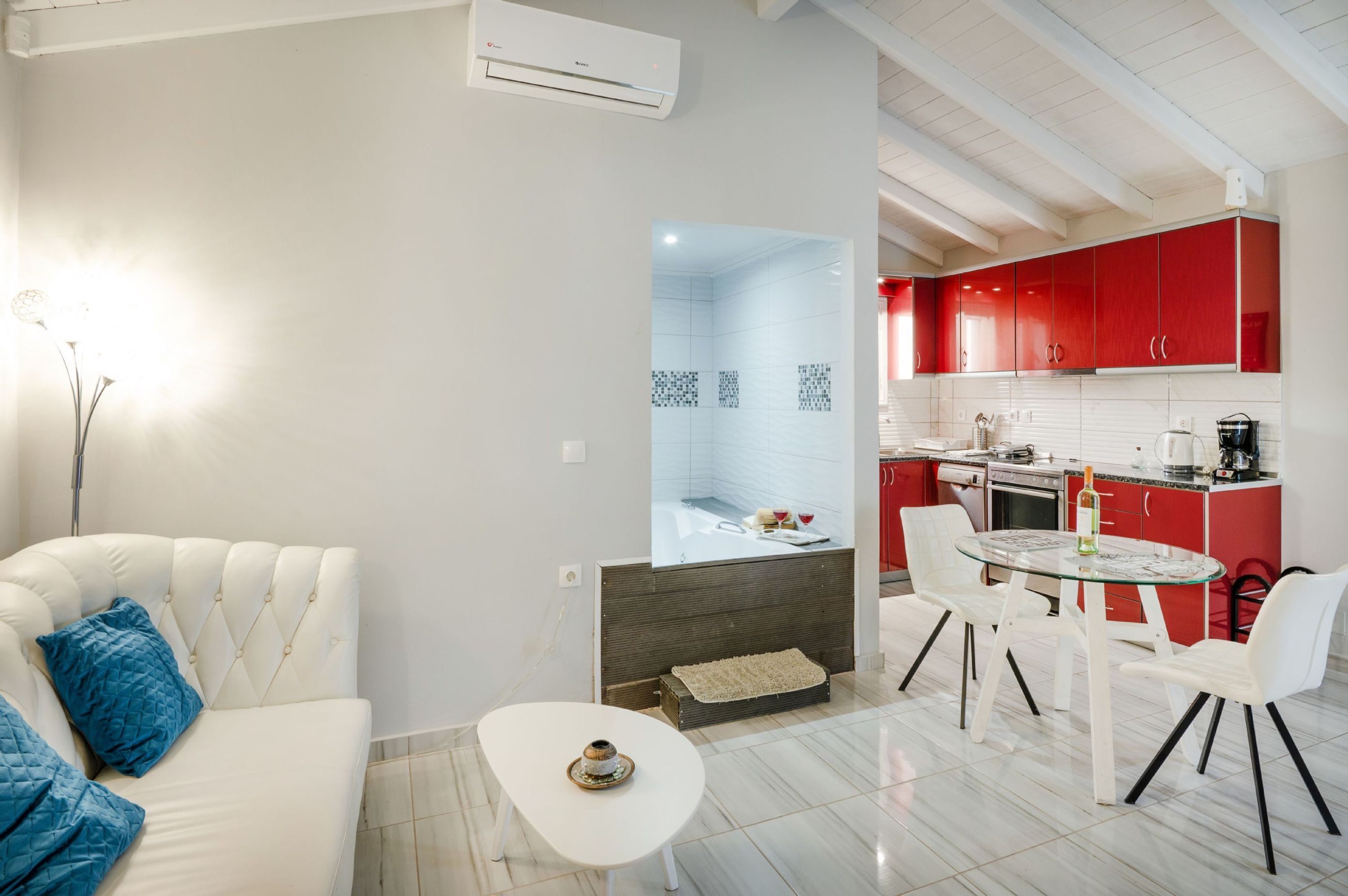 Una sala de estar moderna con un sofá blanco, una mesa de centro de cristal y una cocina roja al fondo.