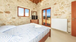 Un dormitorio con paredes de piedra, techo de madera, cama con colcha estampada, cómoda, televisor y vista al mar a través de un ventanal.