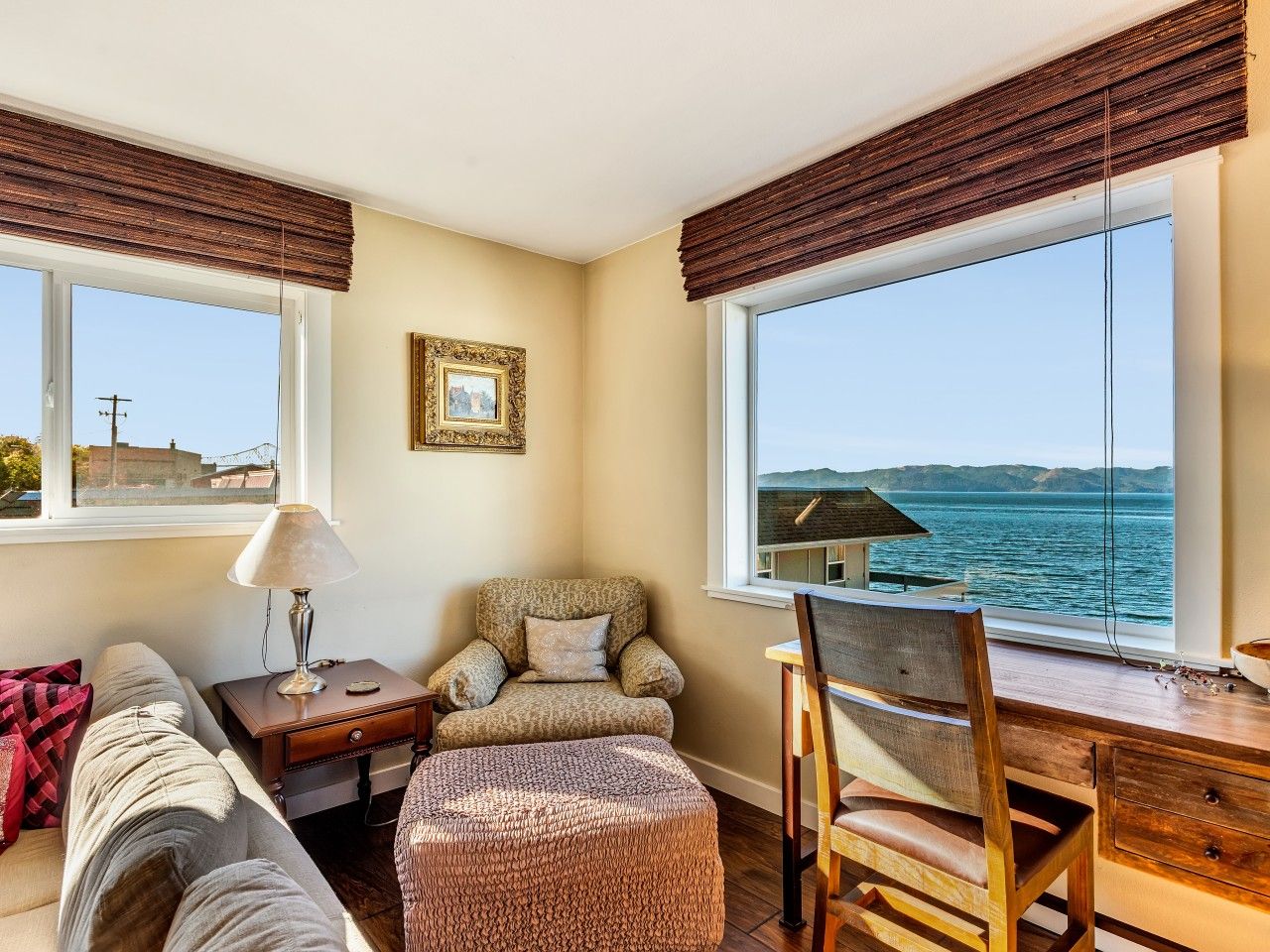 A room with a sofa, armchair, and ottoman, a wooden table, and a view of the sea through large windows.