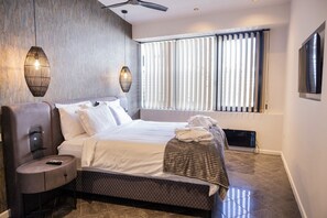Ein modernes Hotelzimmer mit einem großen Bett, einem Nachttisch mit Fernbedienung, einem an der Wand montierten Fernseher und einem großen Fenster mit Jalousien.