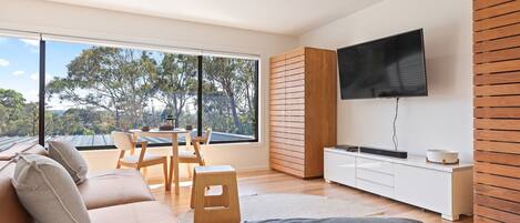 A modern living room with a large window, a flat-screen TV, a sofa, a dining table, and a small stool.
