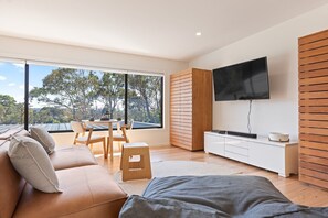 Ein modernes Wohnzimmer mit großem Fenster, Flachbildfernseher, Sofa, Esstisch und einem kleinen Hocker.