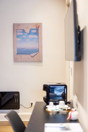 A room with a wooden-framed painting of a sky and clouds, a flat-screen TV mounted on the wall, a desk with a computer and a coffee machine, and a chair.
