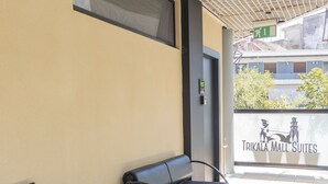 Un espace extérieur aménagé, abrité, avec deux chaises noires et une petite table, un sol carrelé et un mur sur lequel est affiché un panneau indiquant « TRIKALA MALL SUITES ».