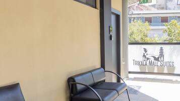 A covered outdoor seating area with two black chairs and a small table, a tiled floor, and a wall with a sign reading "TRIKALA MALL SUITES".