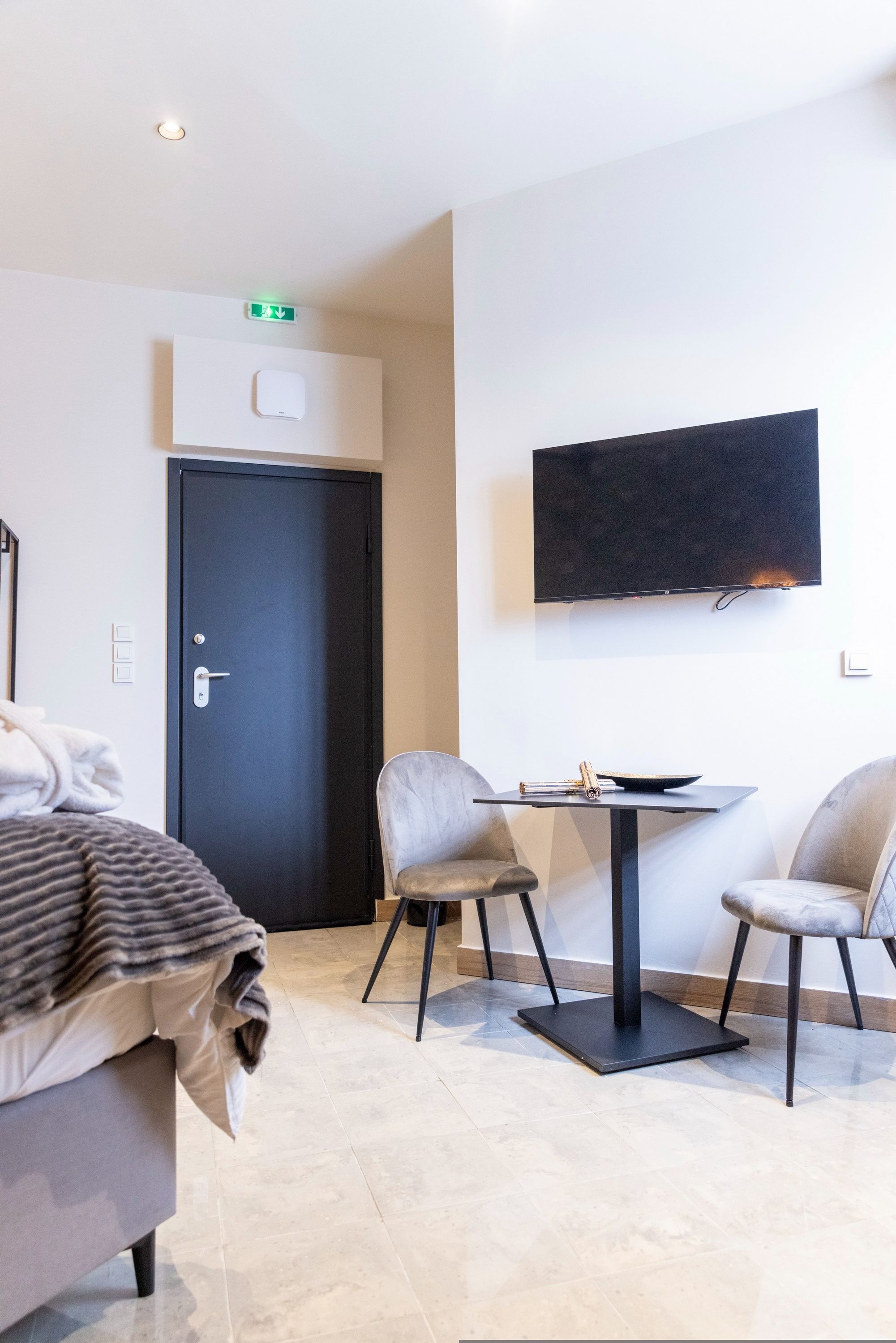 Una moderna camera d'hotel con un letto, una scrivania con due sedie, una TV a schermo piatto montata a parete e una porta con il segnale di uscita di emergenza.