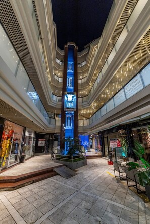 A multi-level atrium with a central illuminated column, glass railings, and various shops on the ground floor. - Trikala Mall Suites 1st Floor Suite Number 10 (Trikala)