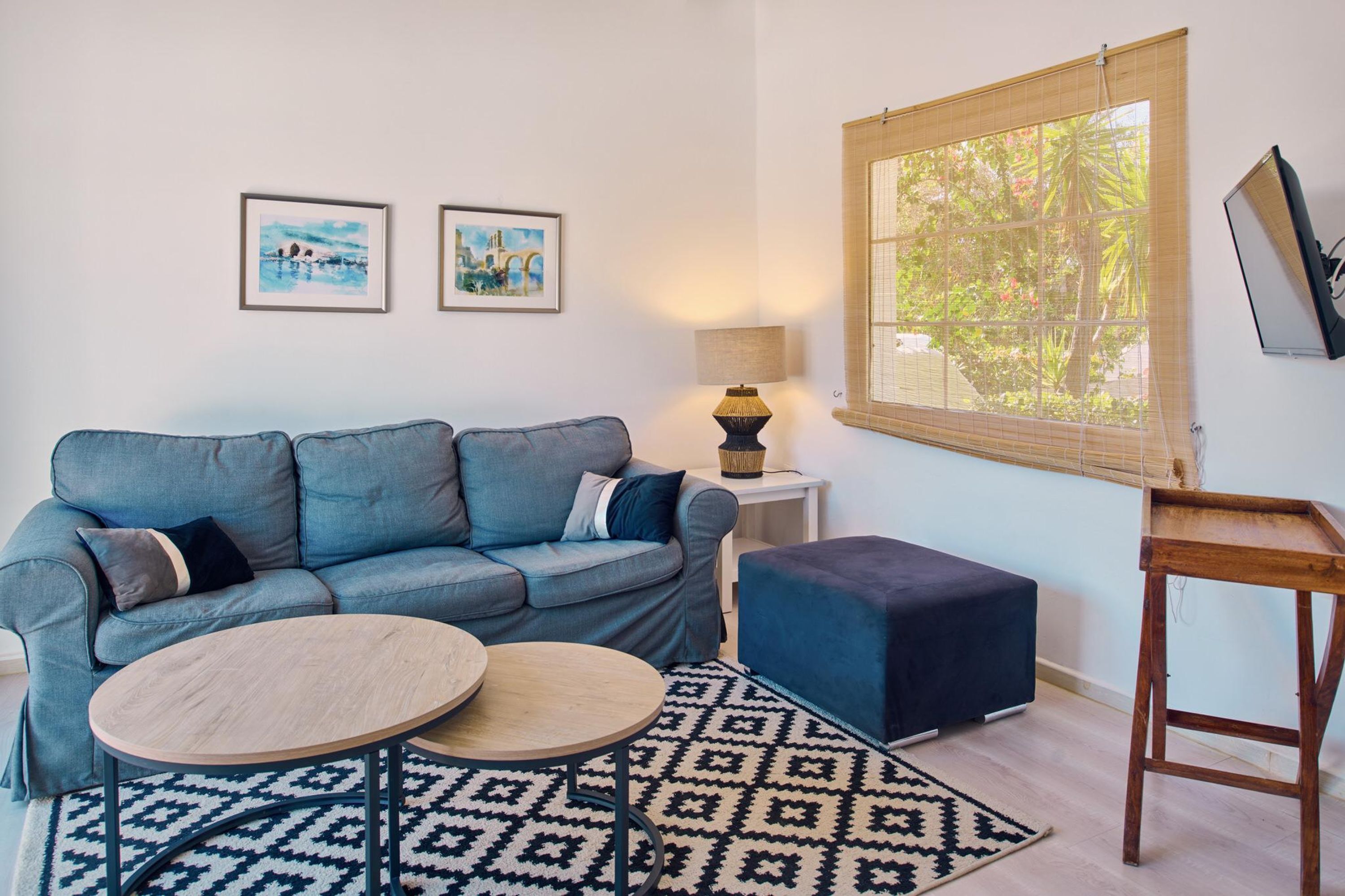 Un salón con un sofá azul, dos mesas redondas de madera, un tapete estampado, una mesita auxiliar con una lámpara y una ventana con vistas a zonas verdes.