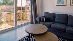 Una sala de estar con un sofá gris, una mesa de centro redonda de madera y un ventanal con vistas a un muro de piedra y zona de estar al aire libre.