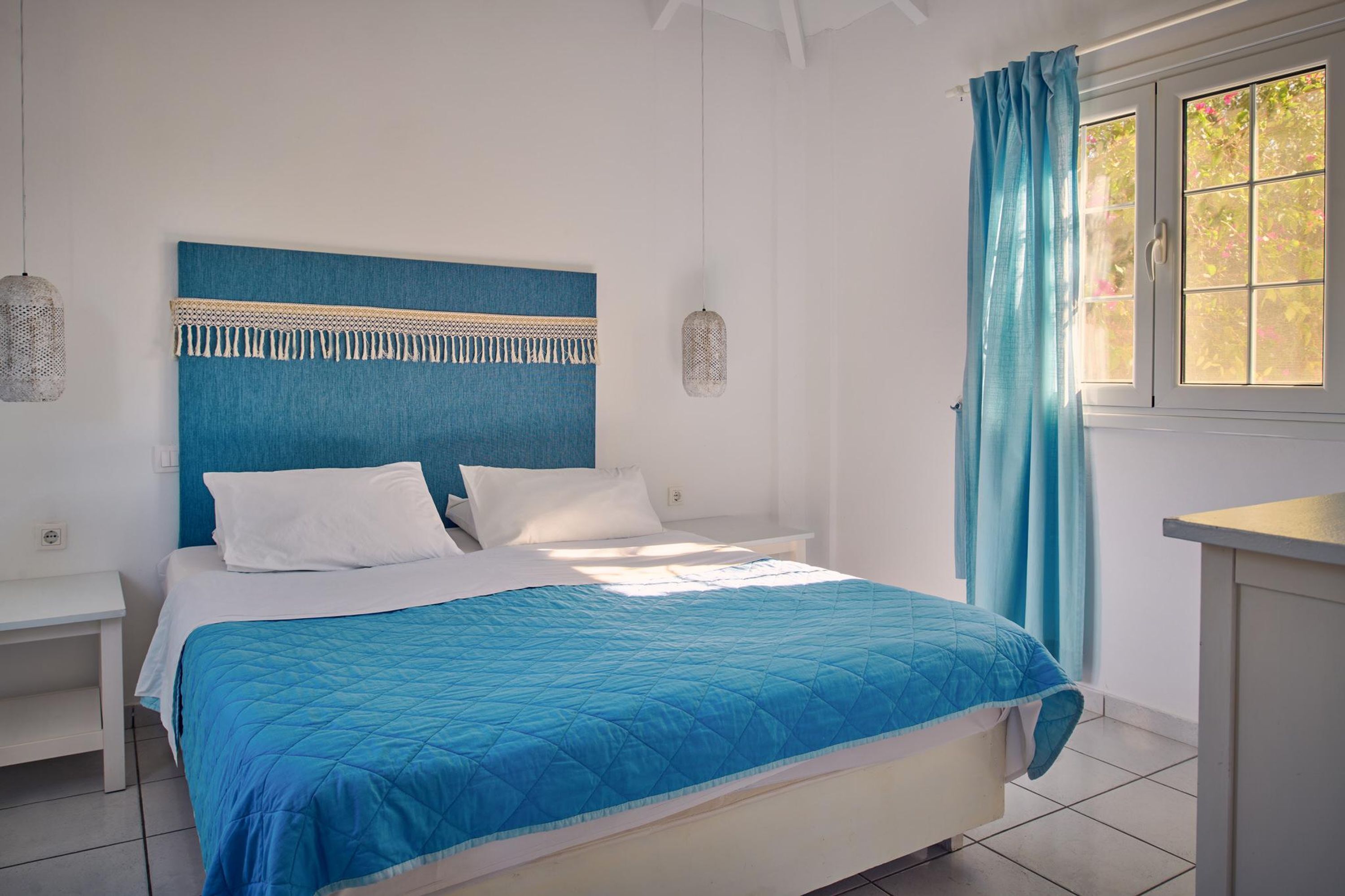 A bedroom with a bed, white nightstands, a blue headboard, and a window with curtains.