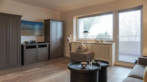 A modern living room with a grey cabinet, a grey sofa, a round coffee table, and a window with blinds. - Fiedler Oase vacation apartment on Sylt - comfort close to the beach (Sylt)
