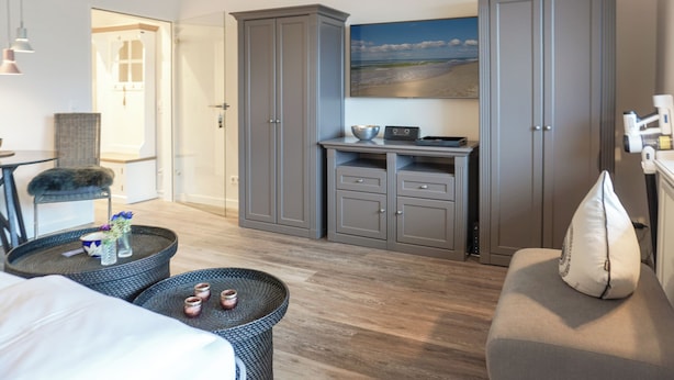 A hotel room with a bed, a seating area with a round table, grey wardrobes, a painting of a beach, and a door leading to another room. - Fiedler Oase vacation apartment on Sylt - comfort close to the beach (Sylt)