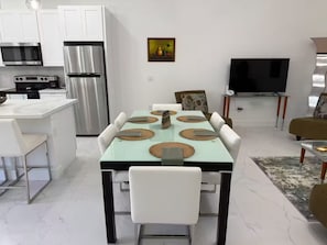 A modern kitchen with white cabinets, a stainless steel refrigerator, and a microwave. There is a dining table with white chairs and a TV in the background. - Chic New Build Duplex /King Suite & Cover Lanai (Punta Gorda)