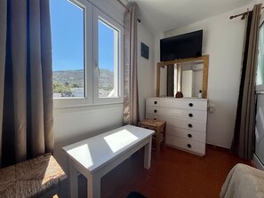 A bedroom with a bed, a desk, a dresser, a TV, and a window with curtains. - Stephany's Skyline Studio (Paros)