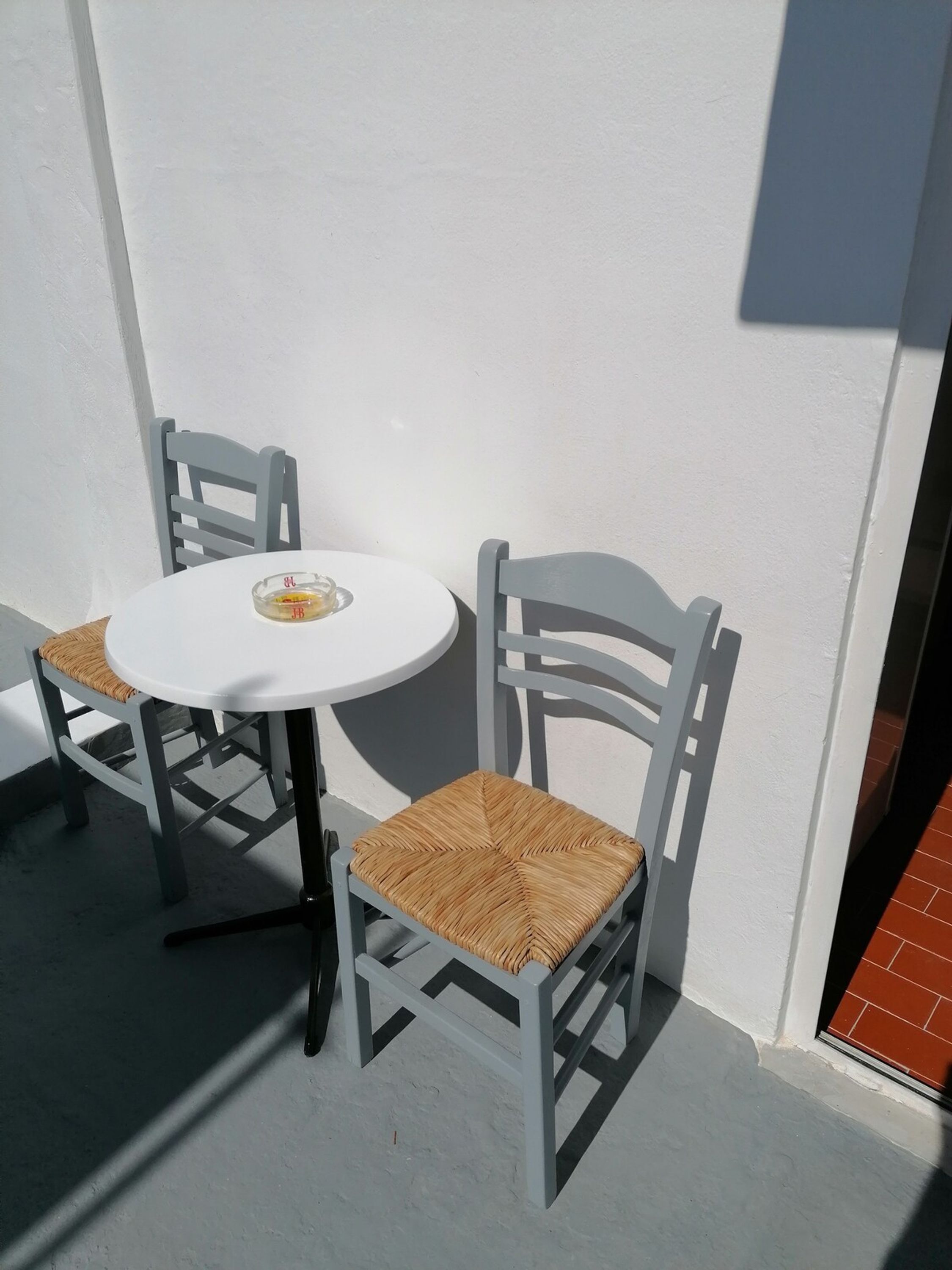 Uma pequena área de estar ao ar livre, com uma mesa redonda e duas cadeiras, uma com assento trançado e outra com assento acolchoado.