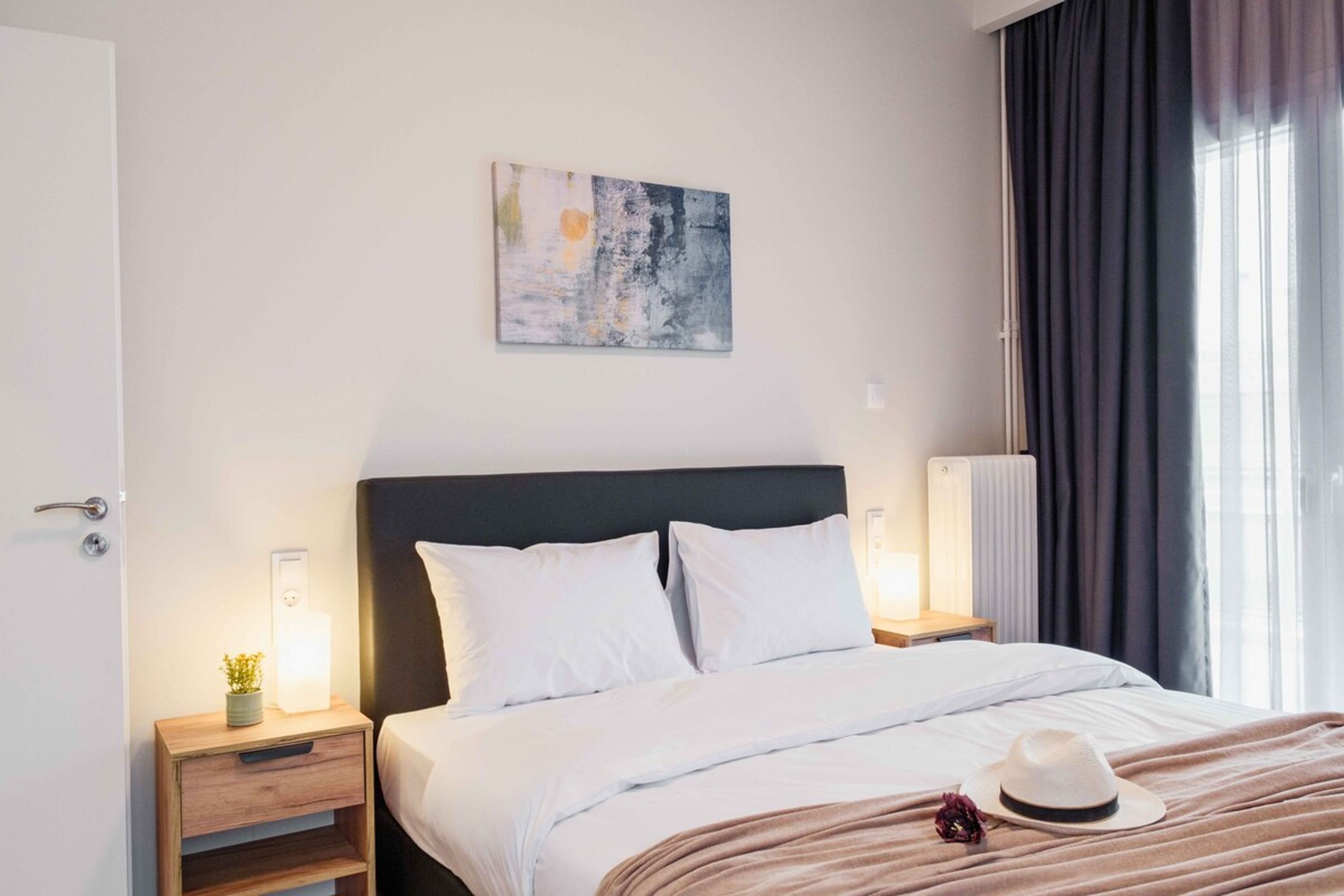 Una habitación de hotel con cama, mesitas de noche, una obra de arte en la pared y una ventana con cortinas.