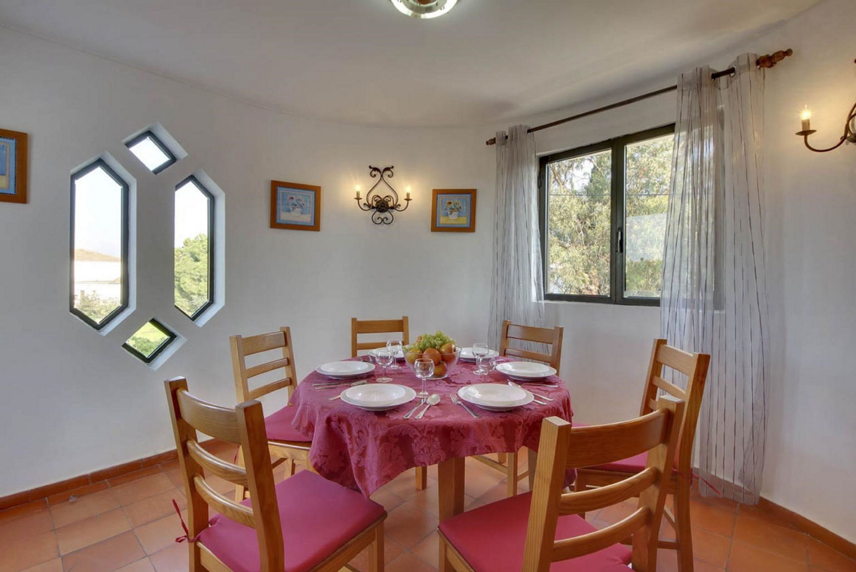 A dining room with a round table set for four, wooden chairs with pink cushions, a red tablecloth, and a chandelier.