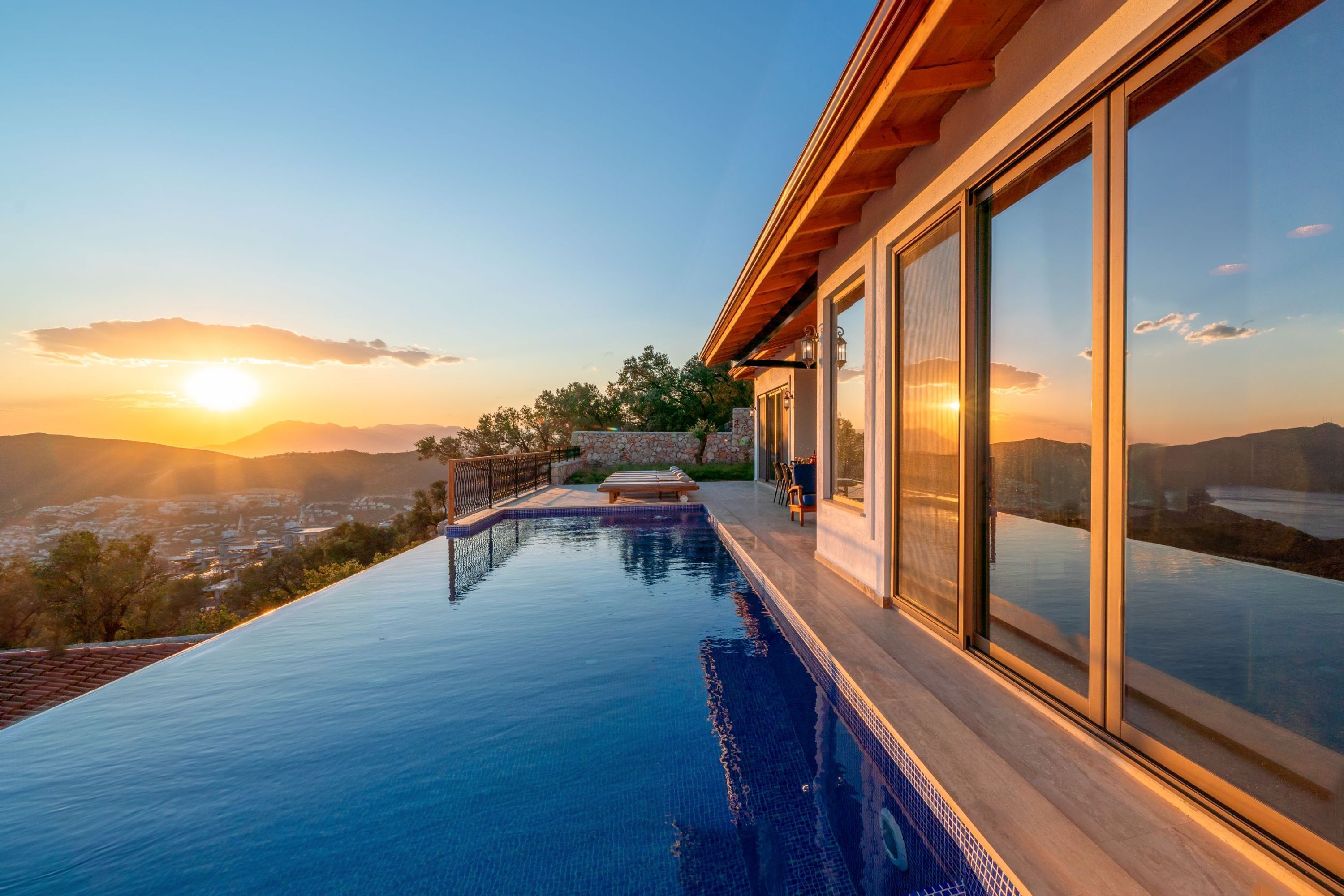 A modern infinity pool with a view of the sunset over mountains, a wooden deck, and large glass doors.