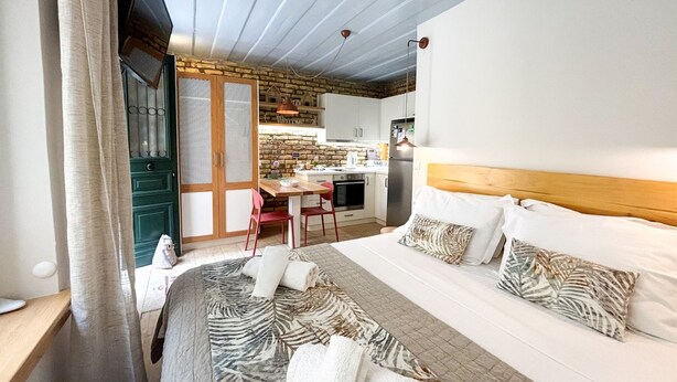 Un dormitorio con una cama grande, cabecera de madera y vista a una cocina con pared de ladrillo y armarios blancos.