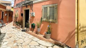 A narrow cobblestone street with a pink building featuring a balcony, potted plants, and a door. - Vilia Port House- Corfu Centre (Kerkira)
