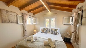 A bedroom with a wooden ceiling, a bed with white bedding and tropical pillows, a window with curtains, and a small round table with a lamp. - Vilia Port House- Corfu Centre (Kerkira)