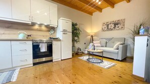 A modern kitchen with white cabinets, a built-in oven, and a refrigerator. There is a grey sofa and a small round table with a lamp in the living area. - Vilia Port House- Corfu Centre (Kerkira)