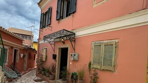 A narrow stone-paved courtyard with a pink building featuring shutters and potted plants. - Vilia Port House- Corfu Centre (Kerkira)