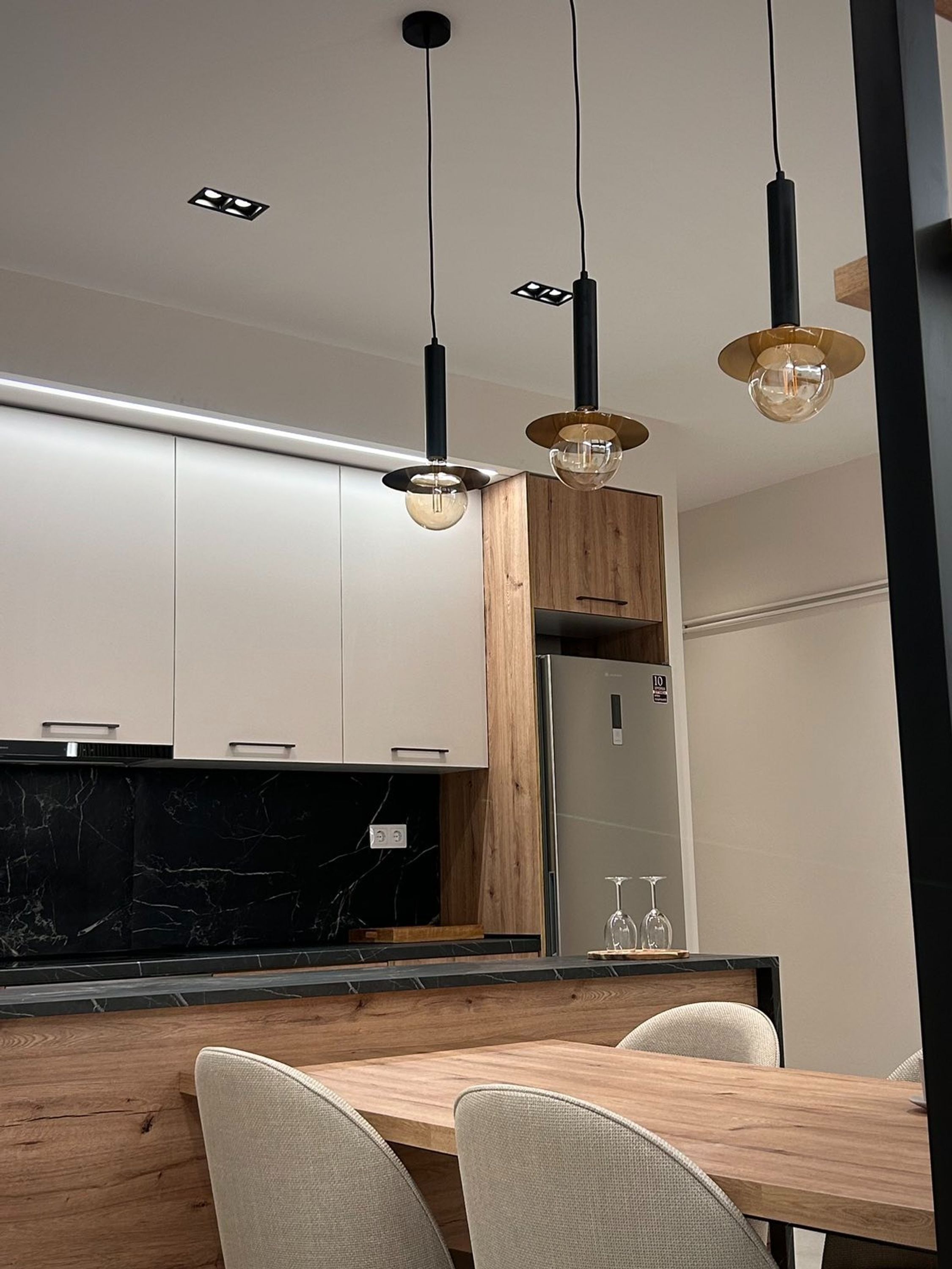 Cozinha moderna com armários brancos, parede de destaque em madeira e bancada de mármore. Há uma área de jantar com mesa e cadeiras de madeira.
