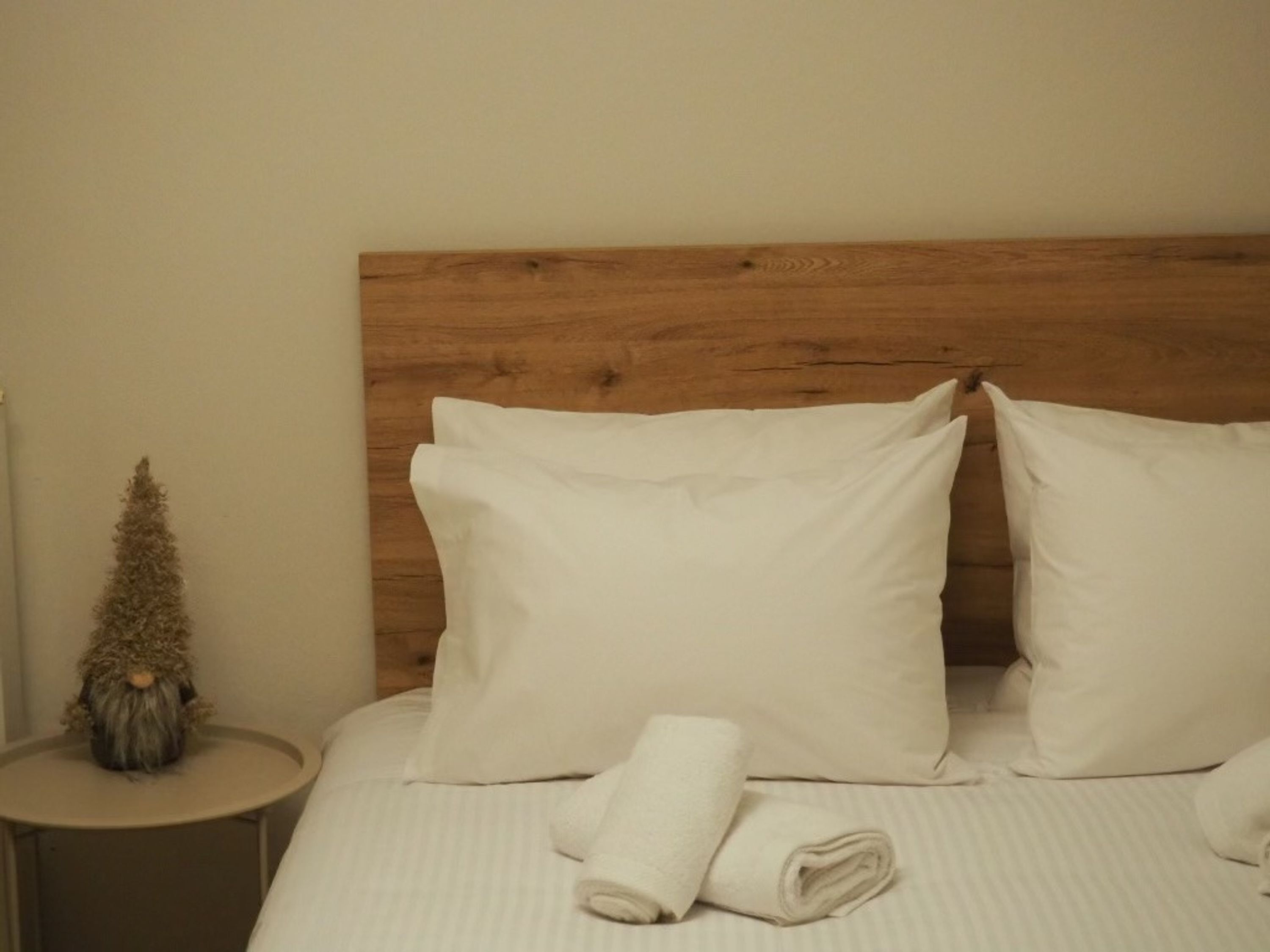 A neatly made bed with white linens and pillows, a small round table with a decorative item, and a wooden headboard.