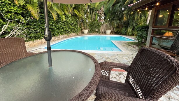 Un espace piscine agrémenté de fauteuils en osier et d’une table en verre, entouré d’une végétation luxuriante et d’une clôture en bois.