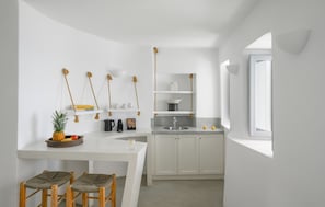 A modern kitchen with white cabinetry, a white countertop, and a small dining area with two stools. There are hanging plants, a coffee machine, and a fruit bowl on the counter. - Executive  Suite with Caldera View & Hot Tub (Santorini)