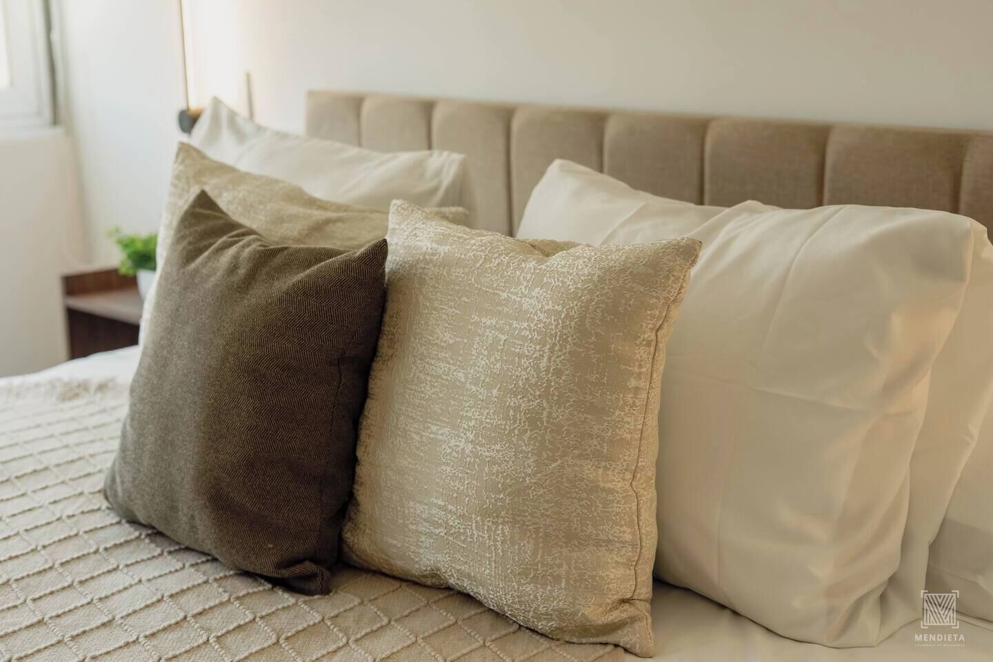 A bed with a beige headboard and three pillows in different shades of beige and brown.