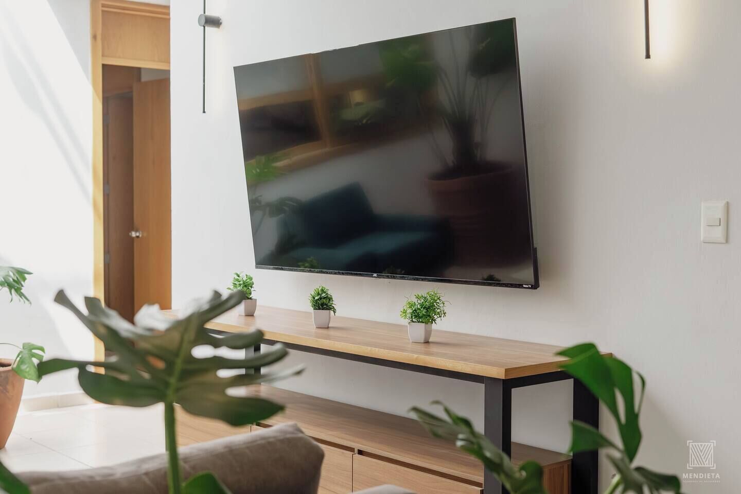 Een moderne woonkamer met een flatscreen televisie aan de muur, een houten salontafel en diverse potplanten.