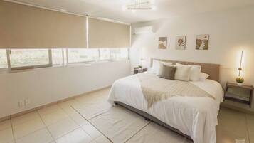 A spacious bedroom with a large bed, a bedside table with a lamp, and a window with blinds.