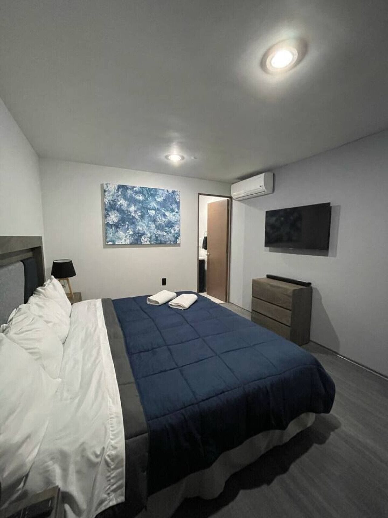 A modern bedroom with a bed, a nightstand, a dresser, a wall-mounted TV, and an air conditioning unit.