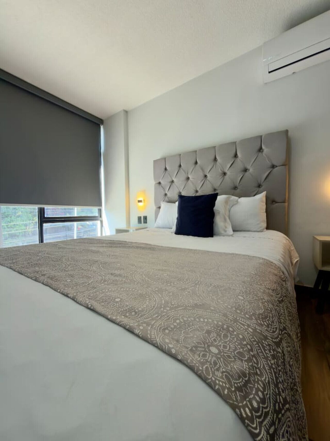 Une chambre d’hôtel avec un grand lit, une tête de lit capitonnée, une couvre-lit à motifs, une table de chevet avec une lampe et une fenêtre donnant sur un beau paysage.