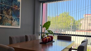 A dining area with a wooden table and chairs, a potted plant, and a large window with vertical blinds. - AR Suites 4: La Calzada (Zamora de Hidalgo)