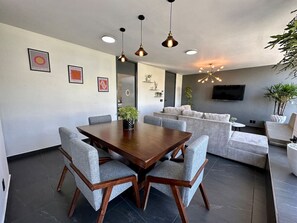 Ein modernes Wohnzimmer mit Essbereich, grauen Polstermöbeln, einem Holztisch und Pendelleuchten.