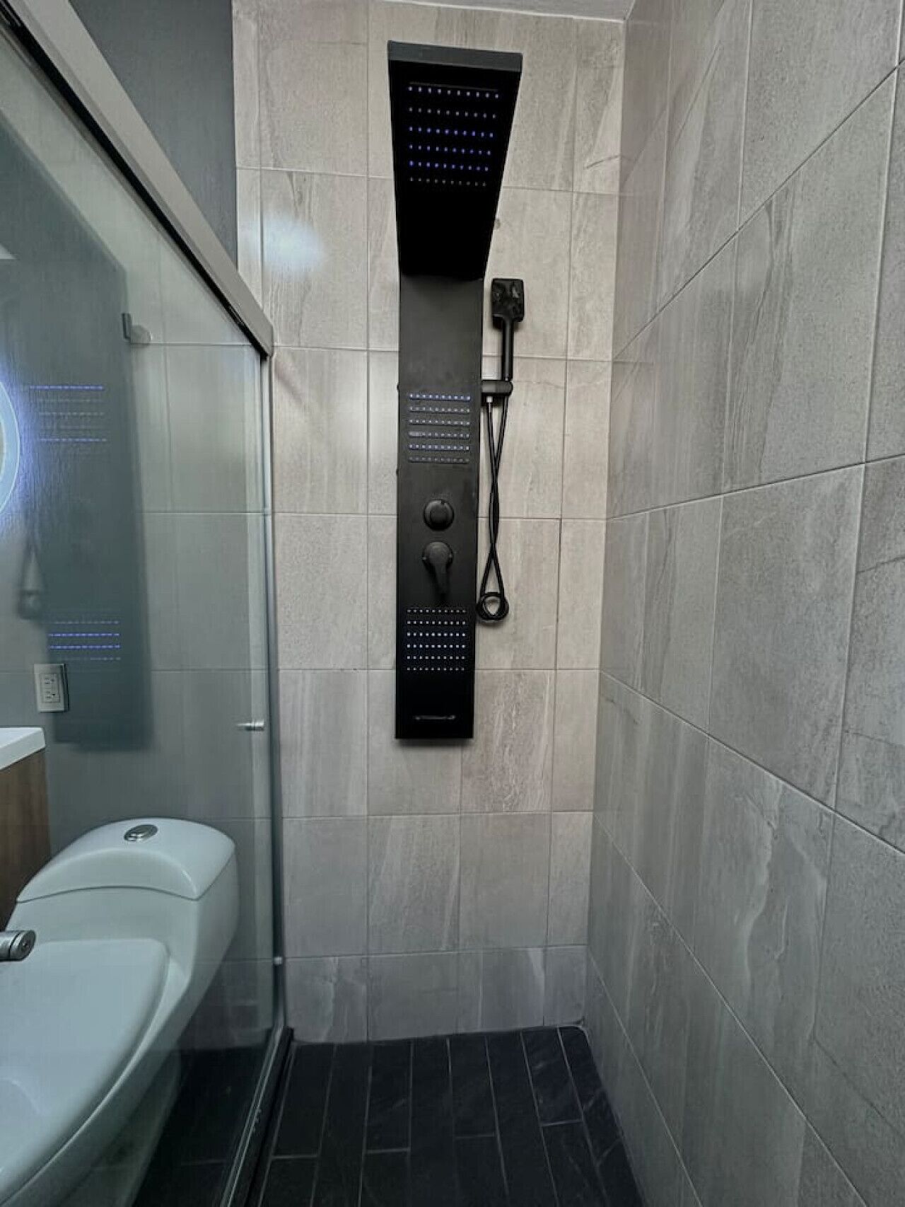Un baño moderno con ducha envuelta en cristal, panel de control negro empotrado en la pared y un inodoro blanco.