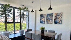 Ein moderner Essbereich mit Holztisch und grau gepolsterten Stühlen, großen Fenstern mit Blumentöpfen und abstrakter Kunst an der Wand.