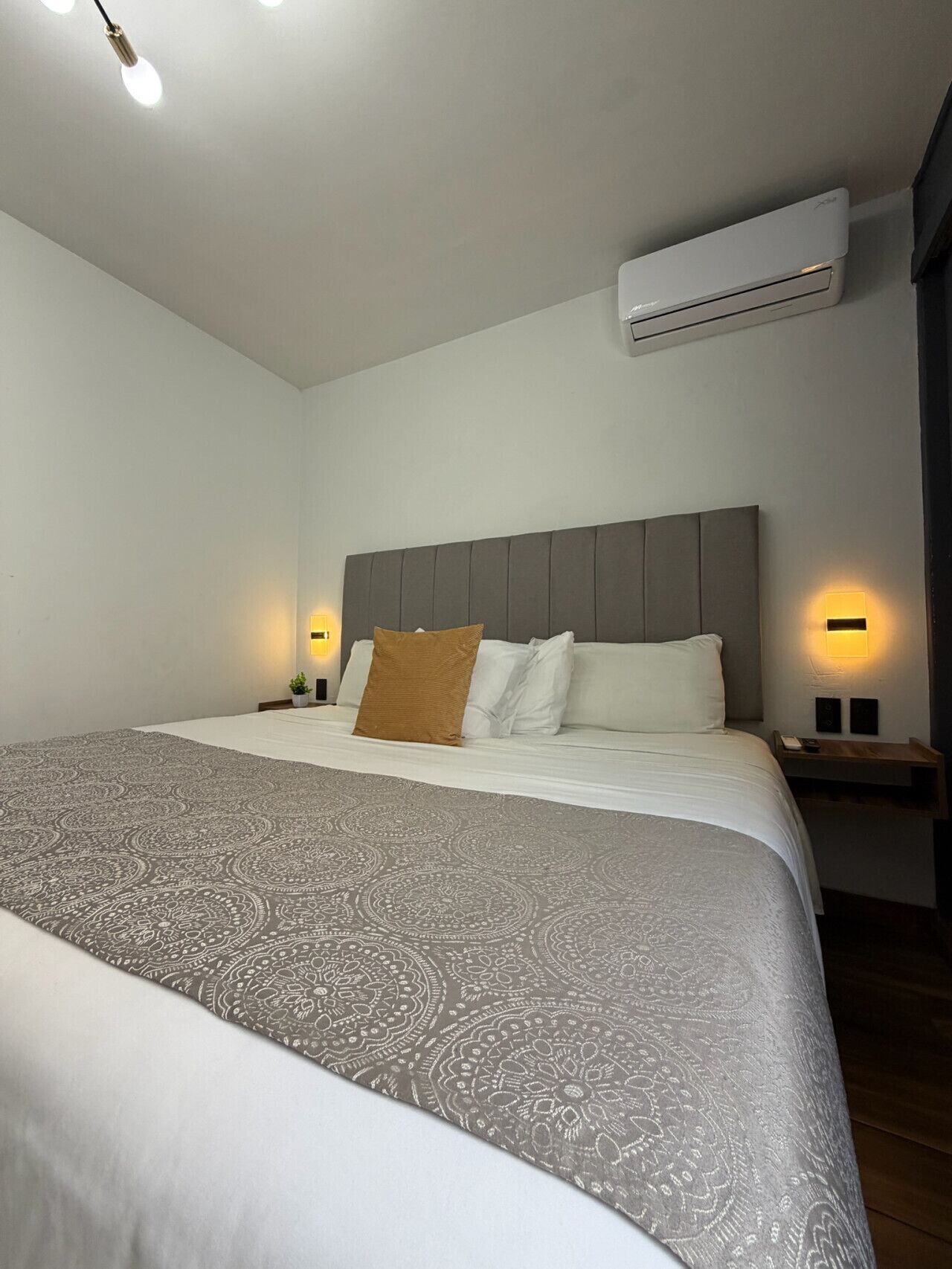 A hotel room with a large bed, a headboard, bedside lamps, an air conditioner, and a nightstand.