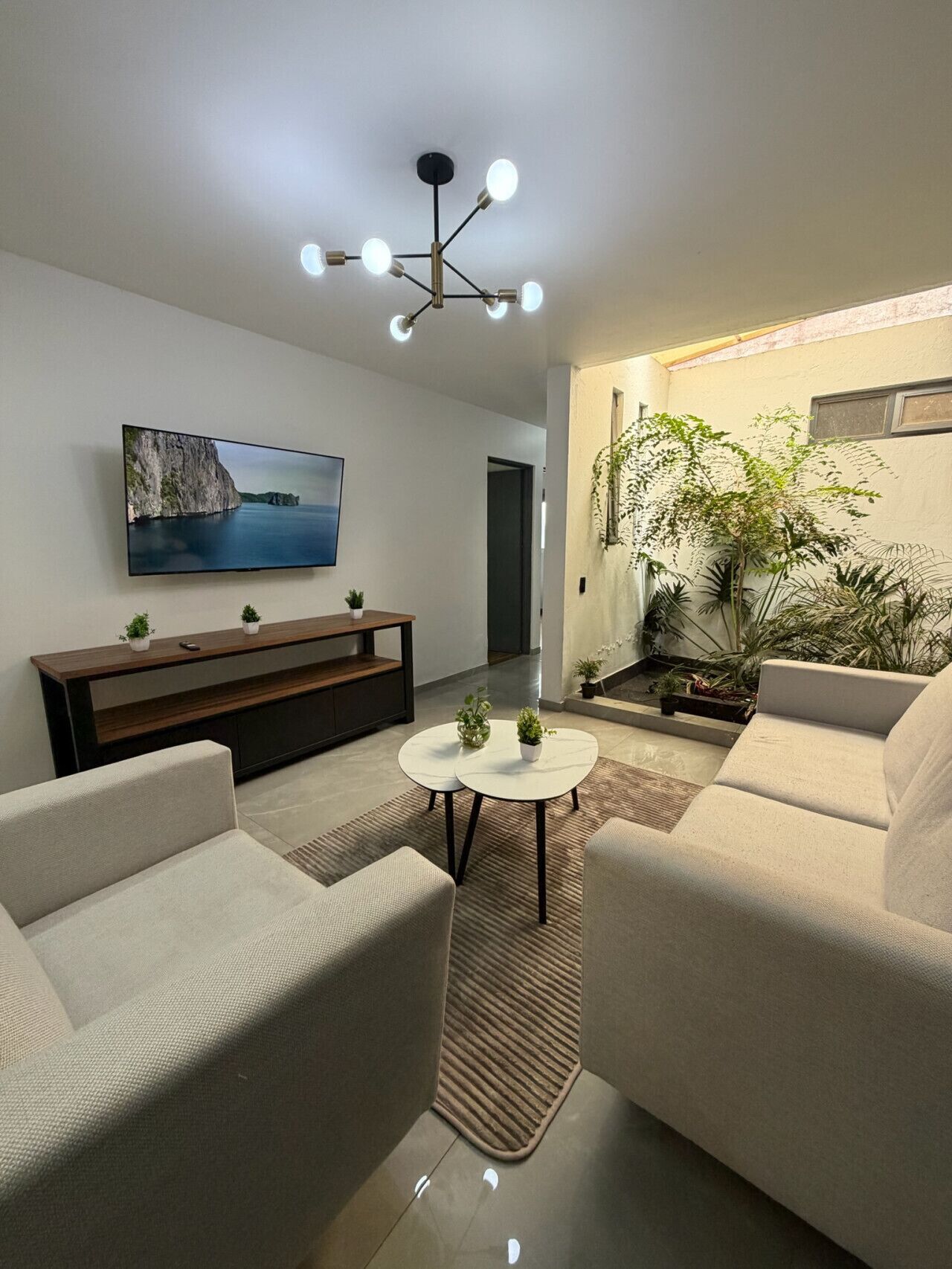 Un salon moderne doté d’un téléviseur à écran plat, d’un meuble de rangement en bois, d’une table basse ronde, de deux canapés, d’un tapis à motifs et d’un luminaire suspendu au plafond.
