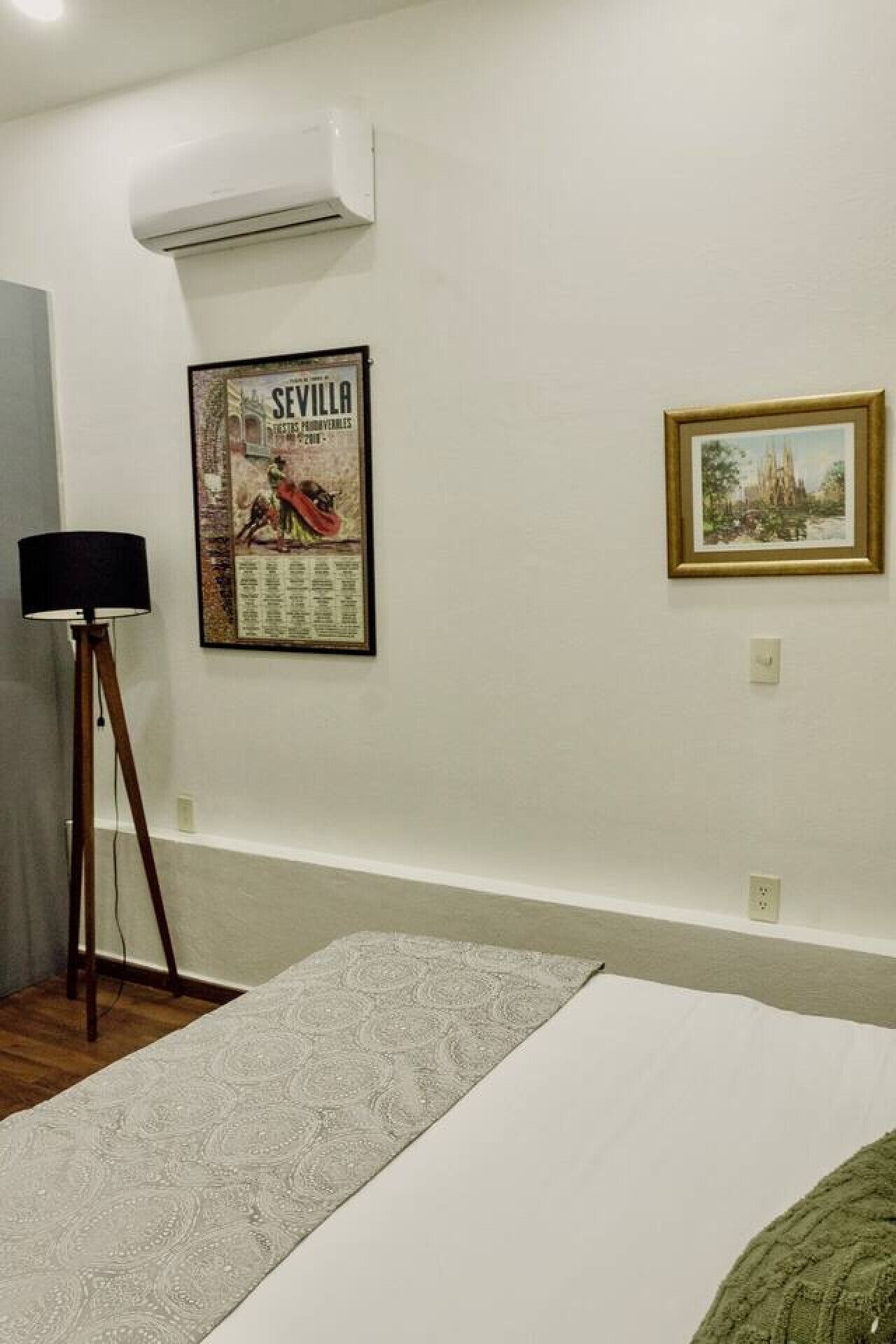 A room with a bed, a floor lamp, an air conditioner, a poster, and a framed picture on the wall.