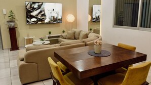 Una sala moderna con un sofá seccional beige, una mesa de comedor de madera con sillas amarillas, un televisor de pantalla plana montado en la pared y una planta en maceta sobre una mesita.