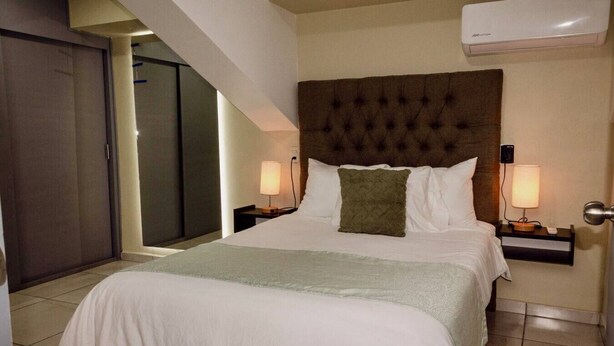 Una habitación de hotel con una cama grande, dos mesitas de noche con lámparas, un cabecero oscuro y un equipo de aire acondicionado en la pared.