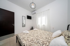 Une chambre à coucher avec un lit, une porte sombre, un téléviseur à écran plat, une petite table de chevet et une fenêtre avec des rideaux.